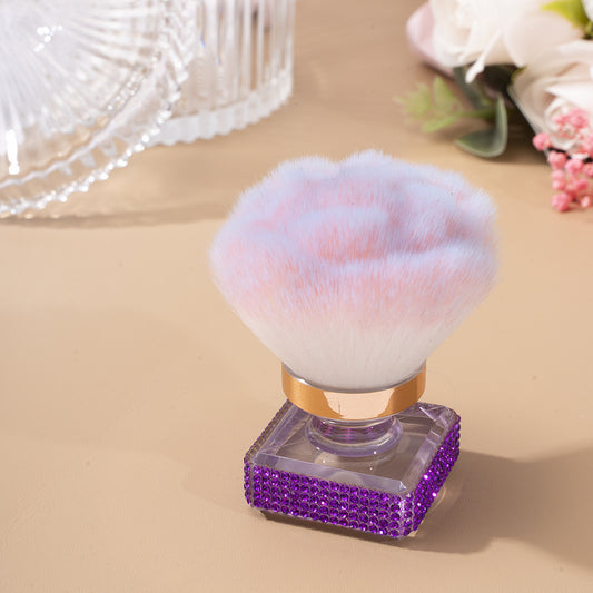 Makeup brush with pink bristle head and purple rhinestone handle on a beige surface.